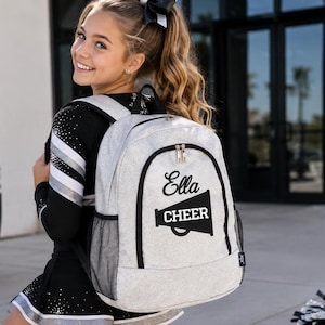 May include: A light gray backpack featuring a black megaphone graphic with the text "Ella" and "CHEER" printed on the front. The bag includes black trim, side mesh pockets, and a top handle, designed for carrying gear with a clear, bold aesthetic.
