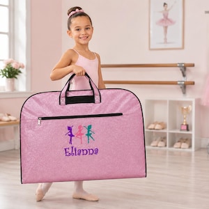 May include: A pink glittery garment bag with black trim and handles. The front features an embroidered graphic of three ballet dancers in purple, pink, and teal, with the name Elianna stitched in purple thread below the figures.