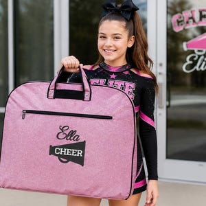 May include: A pink garment bag with black trim and handles, featuring a horizontal zippered pocket. The front is embroidered with the name Ella in script and a megaphone icon with the word CHEER printed in white block letters.