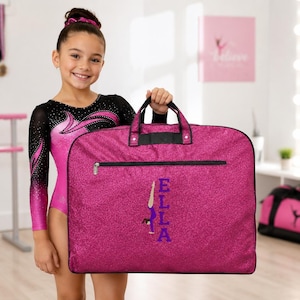 May include: A rectangular, glittery magenta garment bag with black trim and handles. The bag features a horizontal zippered pocket and a vertical graphic of a gymnast in a handstand position next to the purple embroidered name ELLA.