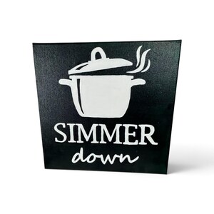 May include: A square black sign featuring a white graphic of a cooking pot with steam rising from the lid. Below the pot, the text reads SIMMER down in white lettering, with the word SIMMER in a bold serif font and down in a script font.