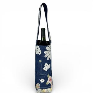May include: A denim wine tote with a dark blue handle and white floral embroidery. The tote has a patchwork design with floral and patterned fabric accents. A green wine bottle is inside the tote.