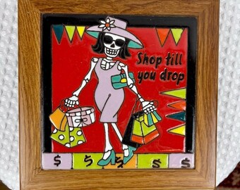 May include: A framed ceramic tile featuring a skeleton woman in a dress and hat, carrying shopping bags. The tile has a red background with the text "Shop till you drop". The frame is made of wood.
