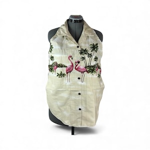 May include: Cream-colored halter top with a tropical flamingo print. The shirt features a button-down front and a collar. The print includes pink flamingos, palm trees, and a landscape scene. The top is displayed on a mannequin.