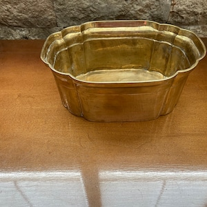May include: A gold-colored brass planter with a decorative, scalloped rim. The planter is empty and sitting on a wooden surface.