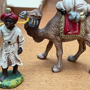 May include: A figurine set featuring a camel and a figure in white robes and a red hat. The camel is brown with a red saddle and white bags. The figure is holding a yellow rope attached to the camel's bridle. The base is green.