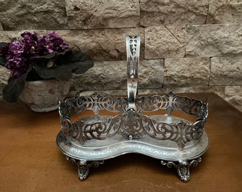May include: Silver plated bottle holder with an ornate design and a handle. The holder has a scalloped base with three feet.
