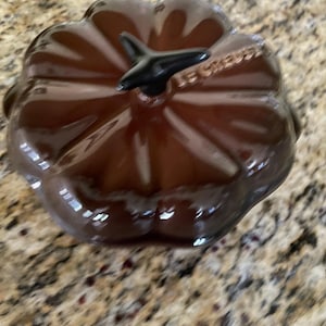 May include: A brown, pumpkin-shaped ceramic dish with a black stem. The dish has a glossy finish and the brand name "LE CREUSET" is visible on the side. The dish is designed for cooking and serving.