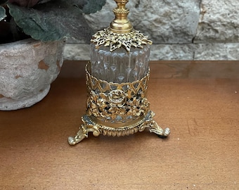 May include: A clear glass perfume bottle with a vertical ribbed texture, encased in an ornate gold-tone metal filigree holder featuring floral motifs and three decorative feet. The stopper is topped with a sculpted gold-tone metal rose.