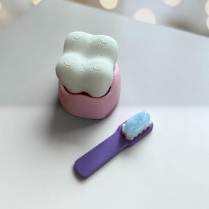 May include: A white, four-lobed tooth-shaped object sits inside a pink, rounded base. Beside it lies a purple toothbrush with a light blue, textured bristle area. Both items feature a distinct, layered 3D-printed texture across their surfaces.