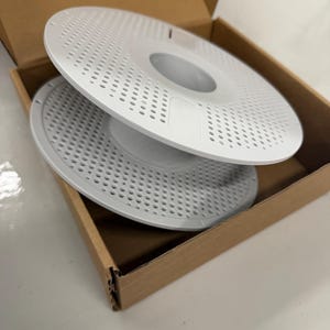 May include: Two white, circular, perforated plastic objects are stacked inside a brown cardboard box. The objects have a central hole and are designed with numerous small, evenly spaced holes across their surface.