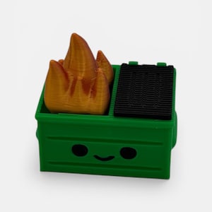 May include: A green dumpster with a smiling face, black lid, and orange flames. The dumpster is a 3D-printed novelty item, likely a desk accessory or a small toy. The flames are a bright orange color, contrasting with the green of the dumpster.