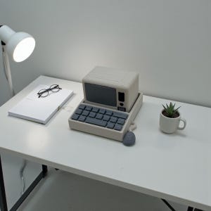 May include: A miniature, retro-style computer model with a gray keyboard, screen, and mouse. A small succulent plant in a white mug and a stack of white paper with glasses are also on the white desk. A white desk lamp is lit.