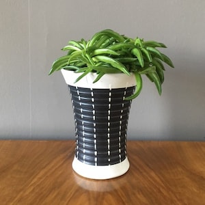 May include: A black and white ceramic planter with a geometric pattern, containing a lush green plant. The planter has a cylindrical form with a white rim and base. The plant's leaves spill over the edge, creating a striking contrast.