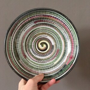May include: A ceramic plate with a spiral pattern. The plate has rings of green, white, red, and yellow, with a black rim. The spiral design is in the centre of the plate. The plate is held by a hand.
