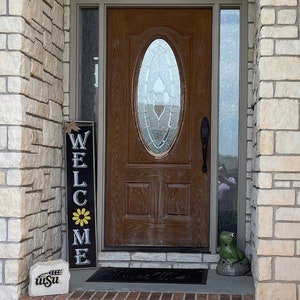 May include: A wooden front door with a glass oval window and a black welcome sign that says "Welcome" with a yellow flower. A small green frog figurine sits on a stone next to the door.