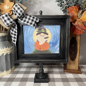 May include: A black framed picture with a Halloween gnome holding a pumpkin. The picture is on a black stand with a square base. The stand is on a white and gray checkered tablecloth.