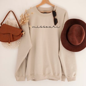 May include: A beige sweatshirt with the word "missouri" written in black script on the front. The sweatshirt is hanging on a wooden hanger and is surrounded by a brown leather bag, a straw hat, and a pair of sunglasses.