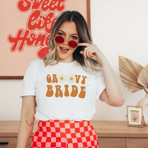 May include: White t-shirt with the text "Groovy Bride" in brown with a daisy graphic. The person wearing the shirt is smiling and wearing red sunglasses.