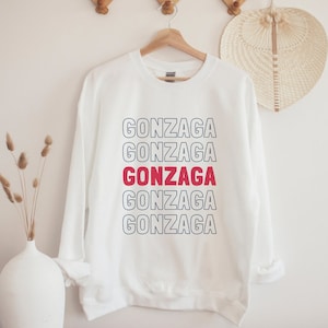 May include: White sweatshirt with the word "GONZAGA" repeated five times in a stacked design. The word "GONZAGA" is in red and outlined in blue in the center of the sweatshirt.