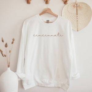 May include: White sweatshirt with the word "cincinnati" written in brown cursive on the front.