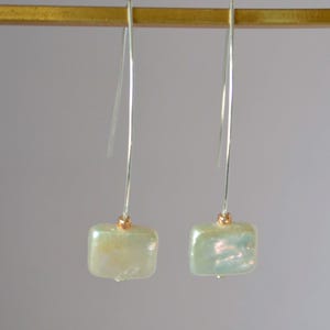 May include: A pair of dangle earrings featuring square, iridescent, light green-yellow beads. The beads are suspended from thin, silver-colored wires with small gold-colored accents. The earrings are displayed against a neutral background.