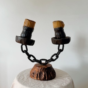 May include: A dual candle holder featuring a carved wooden base, a black metal chain structure, and two dark brown circular platforms. Two thick, amber-coloured wax candles are positioned on the platforms, each with a dark, textured lower section.