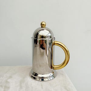 Op de afbeelding: Een roestvrijstalen cafetière met een goudkleurige handgreep en dekselknop. De cilindrische behuizing reflecteert de omgeving. De basis is een iets breder, cirkelvormig ontwerp.
