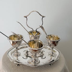 Pode incluir: Um conjunto de porta-ovos prateados com quatro peças de interior dourado, cada uma acompanhada por uma colher a condizer. Os porta-ovos estão dispostos num tabuleiro oval com uma pega central e bordos decorativos recortados.