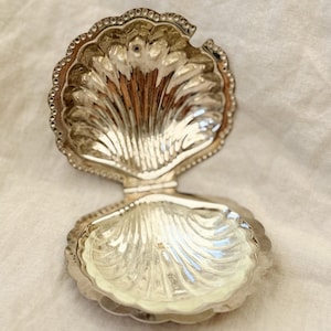 May include: A silver-coloured, hinged seashell-shaped trinket box. The box is open, revealing the interior. The shell design features detailed ridges and a scalloped edge. The box is likely made of metal and sits on a white surface.