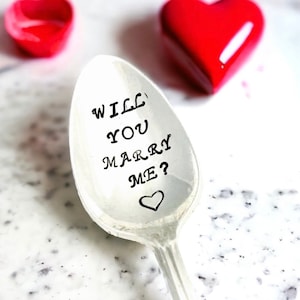Puede incluir: Una cuchara de plata con las palabras "Will You Marry Me?" grabadas en ella. Un pequeño corazón está grabado debajo del texto.