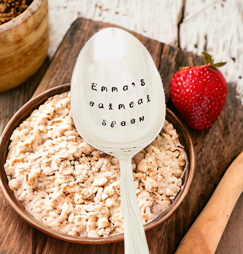 May include: A silver spoon with the text "Emma's oatmeal spoon" is placed over a wooden bowl filled with oatmeal. A fresh red strawberry sits to the right. The spoon is resting on a wooden cutting board.