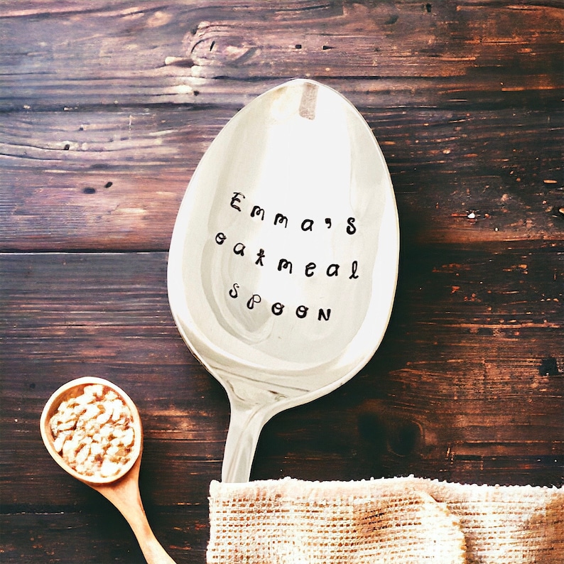 May include: A large, silver spoon with the text "Emma's oatmeal spoon" stamped on the bowl. A small wooden spoon filled with cereal sits to the left. The spoons rest on a dark brown wooden surface.