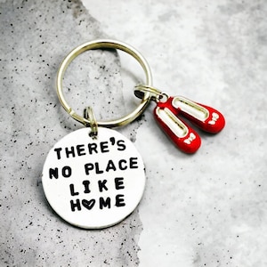 May include: A silver keyring with a round charm that says "There's No Place Like Home" with a heart symbol in place of the "O" in "Home". The keyring also has a charm of two red ruby slippers.