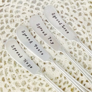 以下が含まれることがあります： 「Spread Hope」、「Spread Peace」、「Spread Joy」、そして「Spread Love」の文字が刻まれた4本の銀色のバターナイフ。ナイフは装飾的なハンドルを持ち、白いかぎ針編みの表面に配置されています。