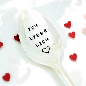 May include: A silver spoon with the words "Ich Liebe Dich" and a heart engraved on it. The spoon is surrounded by small red hearts.