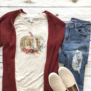 Puede incluir: Una camiseta beige con una calabaza de estampado de leopardo y un diseño floral. La camiseta se combina con una chaqueta de punto marrón, vaqueros azules y zapatos beige de deslizamiento.