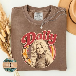 May include: A brown crewneck t-shirt featuring a graphic print of a woman with voluminous blonde hair. The text Dolly is arched above the portrait in red letters. The design includes concentric circles in yellow and orange with scattered stars.