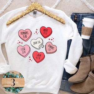 Puede incluir: Sudadera blanca con un gráfico de corazones de caramelo rosa y rojo con versículos bíblicos impresos en ellos. La sudadera está en una percha con un par de jeans y botas marrones.