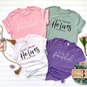 May include: Four t-shirts with Easter-themed messages. The shirts are in pink, white, green, and purple. The messages on the shirts are "It is finished", "Plot Twist He Lives Luke 24:6", and "It is finished".