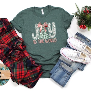 May include: A green t-shirt with the words "JOY TO THE WORLD" in a festive design, alongside a red and black plaid shirt, blue jeans, and white trainers. A small Christmas arrangement is also present.