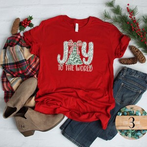 Puede incluir: Una camiseta roja con las palabras "JOY TO THE WORLD" en un diseño festivo. La palabra "JOY" está en un patrón de cuadros, y la palabra "TO" está en un diseño de árbol de Navidad verde. La camiseta se combina con vaqueros azules y botas marrones.