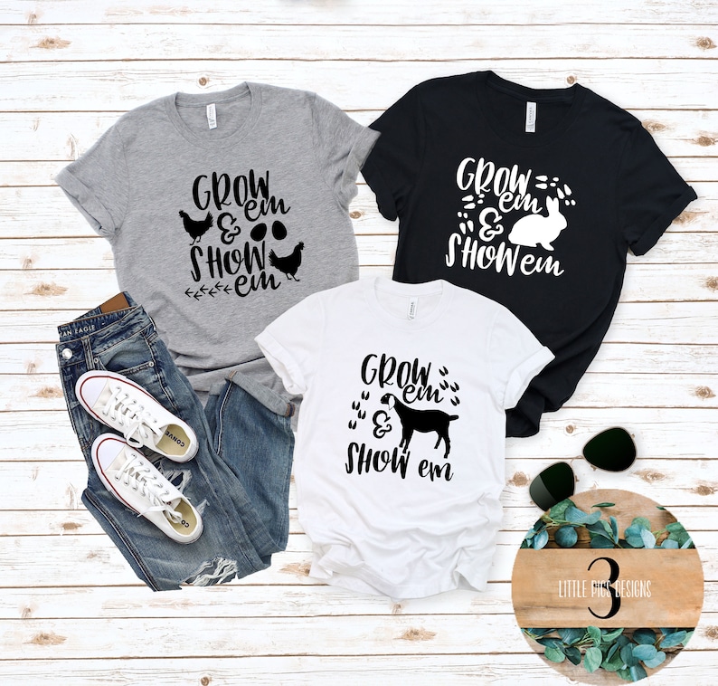 May include: Three graphic t-shirts in gray, black, and white, each featuring the text "Grow 'em & Show 'em" with different animal illustrations: chickens, a rabbit, and a goat. The shirts are displayed with jeans and white sneakers.
