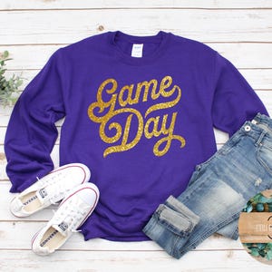 May include: A purple sweatshirt with the text "Game Day" in gold glitter lettering. The sweatshirt is paired with blue jeans and white sneakers. The image is on a white wooden background.