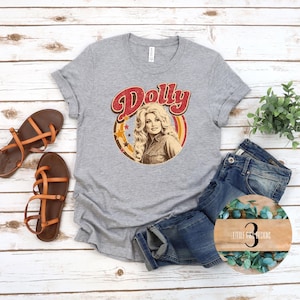 Puede incluir: Una camiseta gris jaspeado con un gráfico circular que muestra el retrato de una mujer rubia, la palabra Dolly en letra roja y estrellas amarillas. La camiseta se presenta con sandalias de tiras marrones y vaqueros azules.