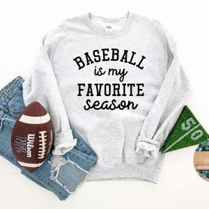 May include: A light gray sweatshirt with the text "Baseball is my favorite season" in black. A brown and white football, blue jeans, and a green field goal marker are also in the image.