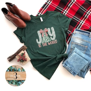 May include: A dark green t-shirt with the words "JOY TO THE WORLD" in a festive design, featuring a plaid bow and Christmas tree. The shirt is paired with blue jeans, brown boots, and a red and black tartan scarf.