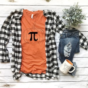 May include: Orange t-shirt with a black pumpkin design featuring the mathematical symbol pi. The shirt is laid out on a white wooden surface with a black and white checked shirt, blue jeans, and a white mug with the text "Pumpkin Spice" in black.