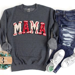 May include: A dark gray sweatshirt with the word "MAMA" in a baseball-themed design. The letters are white with red accents, resembling baseball stitching. The sweatshirt is paired with blue jeans and gray sneakers.