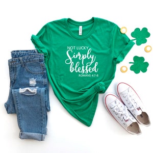 May include: Green t-shirt with white text that reads "Not Lucky Simply Blessed Romans 4:7-8". The shirt is paired with blue jeans and white trainers. There are green shamrocks and gold coins on the white background.